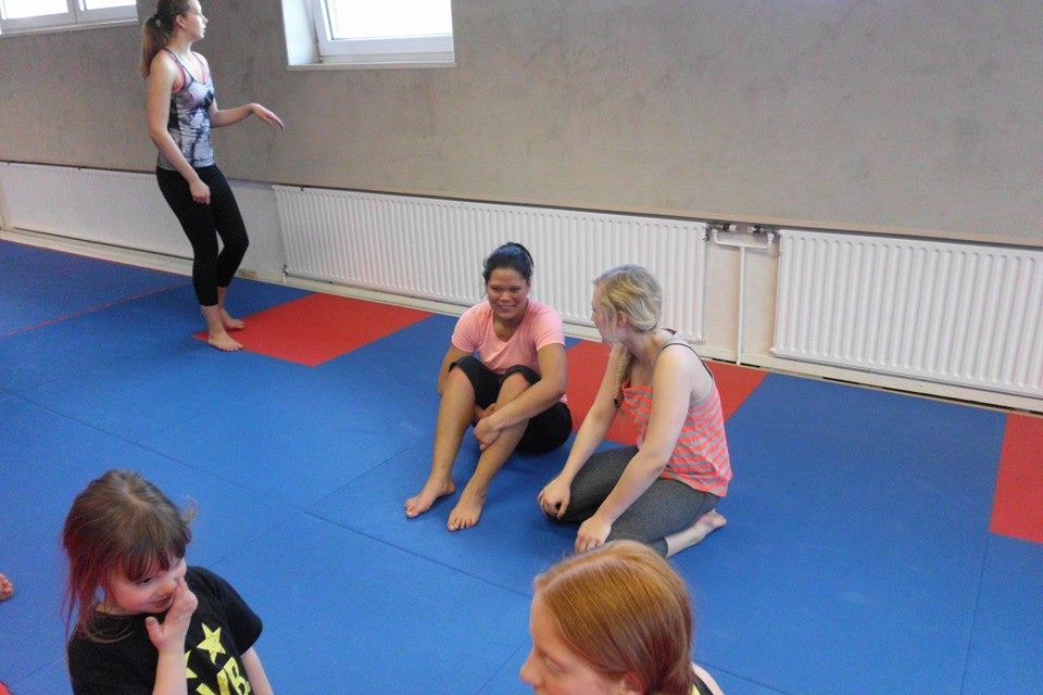 Drei Frauen sitzen auf einer blauen Matte, w&auml;hrend eine weitere im Hintergrund steht. Atmosph&auml;re der Entspannung.