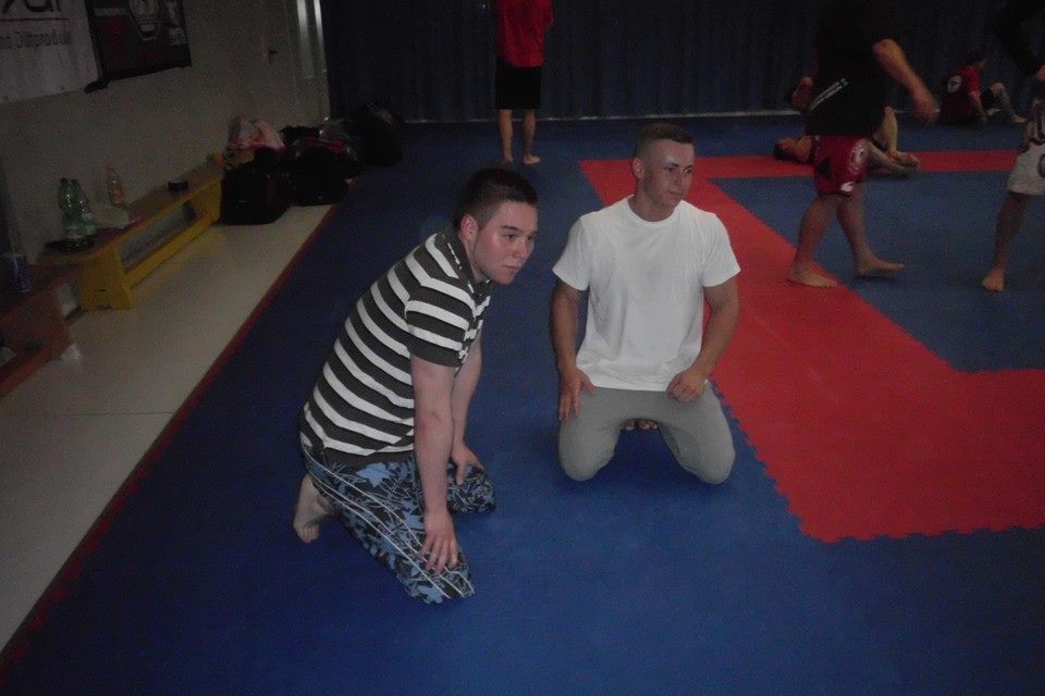 Zwei Personen knien auf einer blauen Trainingsmatte, w&auml;hrend im Hintergrund andere K&auml;mpfer aktiv sind.