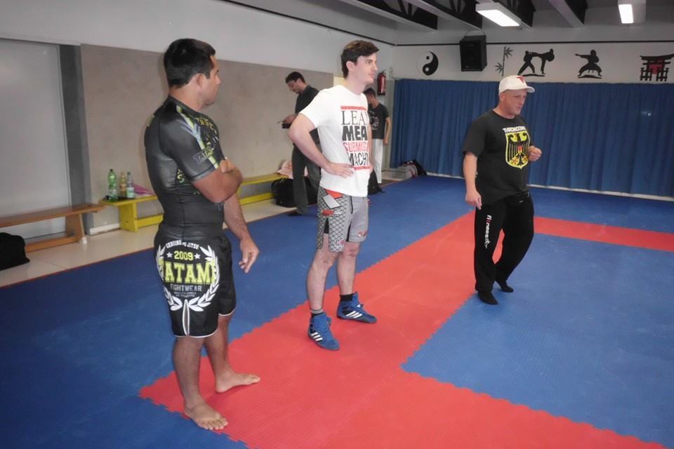 Drei M&auml;nner stehen auf einer Trainingsmatte in einem Kampfsportstudio, bereit f&uuml;r eine Unterrichtseinheit.