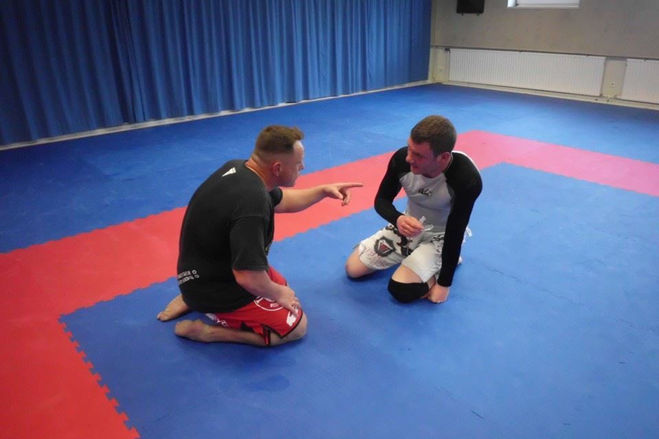 Zwei Kampfsportler sitzen auf einer Matte in einem Trainingsraum, der mit roten und blauen Fl&auml;chen markiert ist.