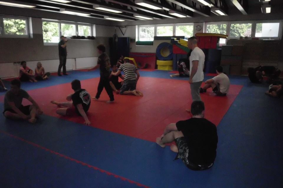 Gruppe von Personen trainiert in einem Sportraum auf blauen und roten Matten, mit verschiedenen Trainingshaltungen.