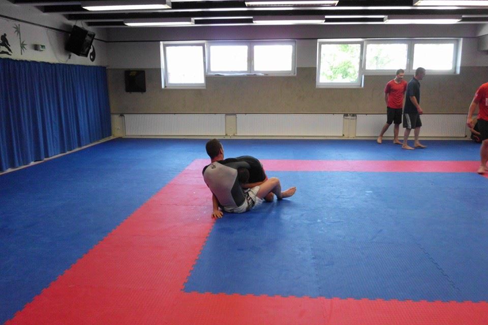 Zwei Sportler im Bodenkampf auf einer blau-roten Mattenfl&auml;che in einem Trainingsraum f&uuml;r Kampfsport.