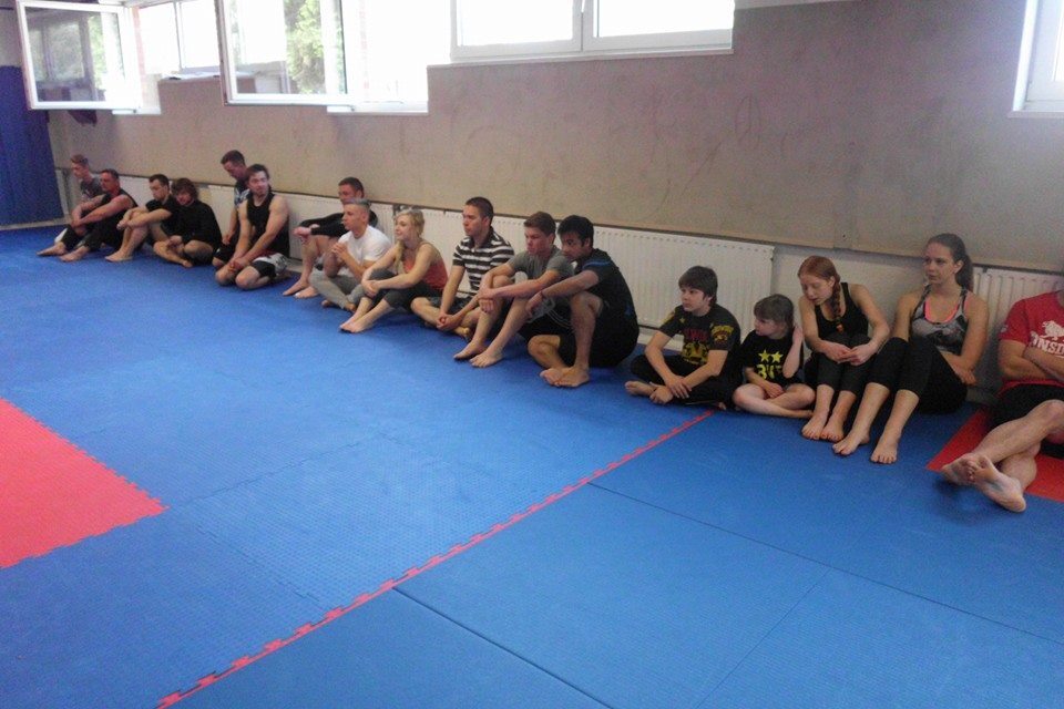 Gruppe von Personen, die auf blauen Matten in einem Raum sitzen, w&auml;hrend sie aufmerksam einem Training zusehen.