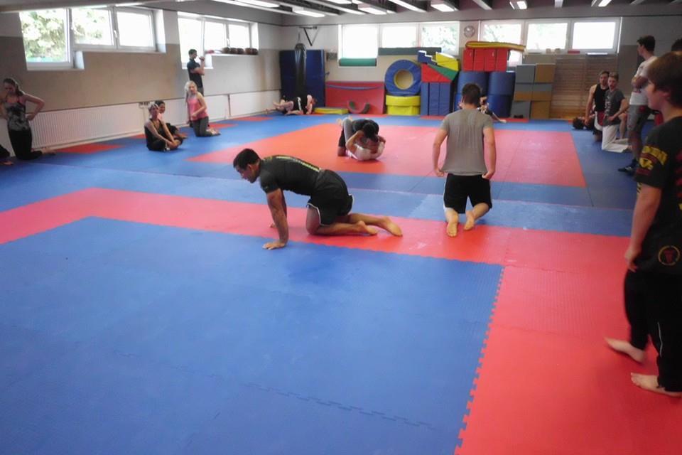 Sportliche Trainingseinheit in einem &Uuml;bungsraum mit Matten, mehreren Personen auf dem Boden und an der Wand.