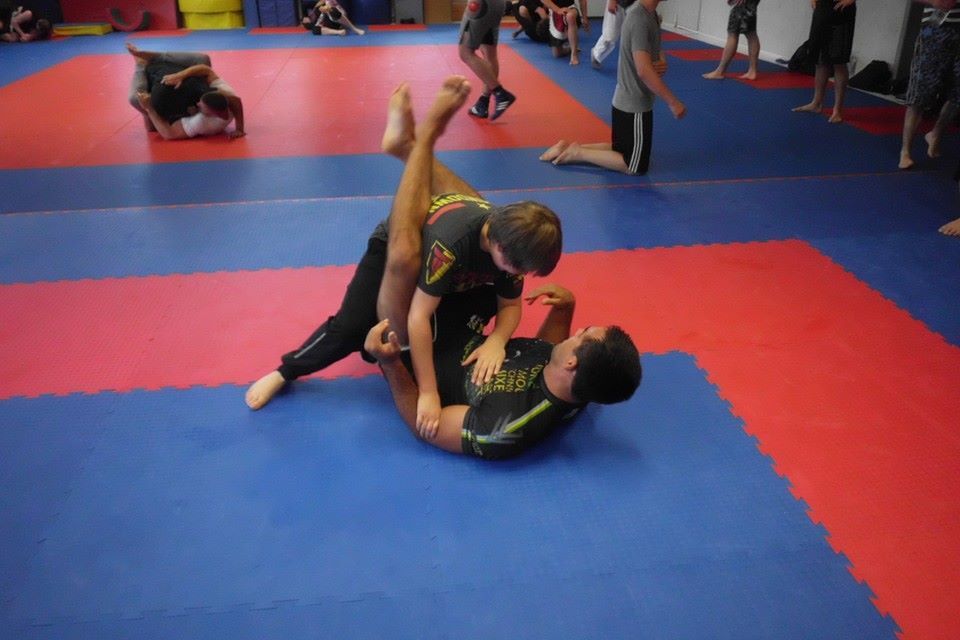 Zwei K&auml;mpfer in Sportkleidung &uuml;ben Bodentechniken auf einer blau-roten Mattenfl&auml;che in einem Trainingsraum.