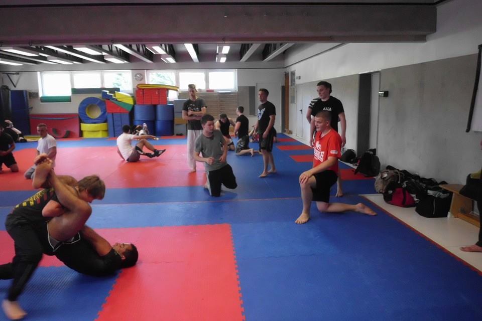 Gruppe von Menschen trainiert auf einer blauen Matte; einige &uuml;ben Grappling-Techniken, andere beobachten.