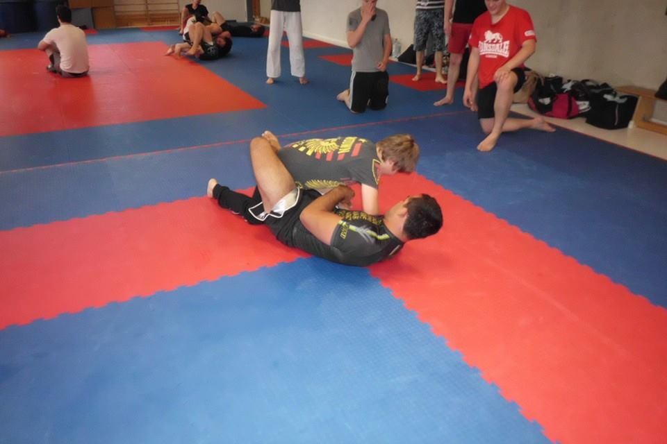 Zwei K&auml;mpfer &uuml;ben auf einer blauen und roten Matte, w&auml;hrend Zuschauer im Hintergrund zuschauen.