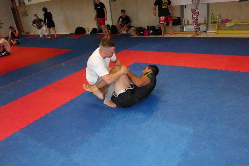 Zwei K&auml;mpfer &uuml;ben Grappling-Techniken auf einer blauen und roten Trainingsmatte in einem Sportraum.