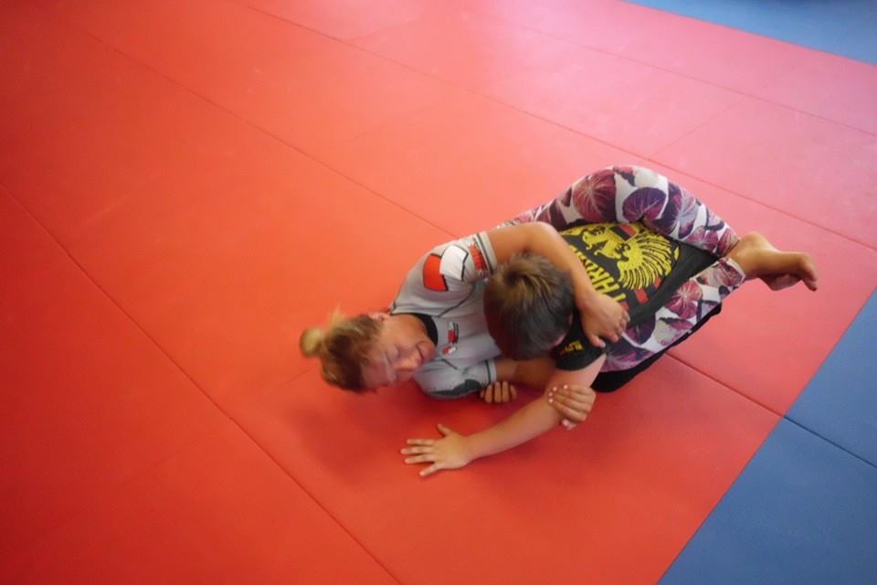 Zwei Personen trainieren Jiu-Jitsu auf einer roten Matte, eine im Oberk&ouml;rperhalt, die andere darunter geklemmt.