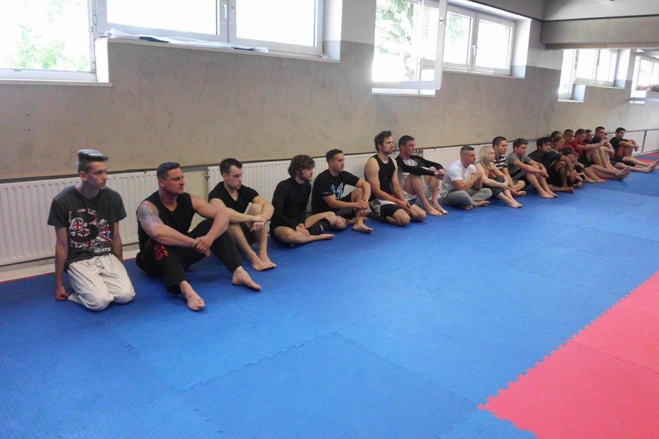 Gruppe von M&auml;nnern in Sportswear sitzt auf einer blauen Matte in einem Trainingsraum. Fenster mit Tageslicht im Hintergrund.