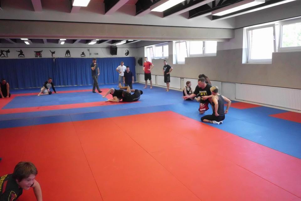 Gruppentraining auf einer Matte mit verschiedenen K&auml;mpfern in einem Trainingsraum mit blauen und roten Bereichen.