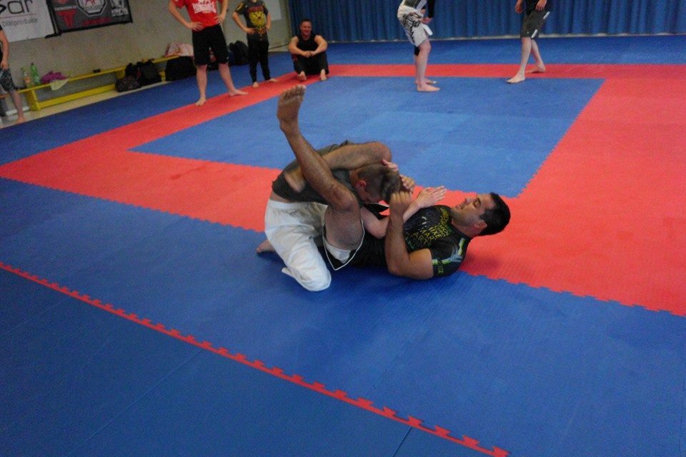 Zwei Sportler im Kampftraining auf blauen und roten Matten, einer im Halbmondposition, der andere am Boden.