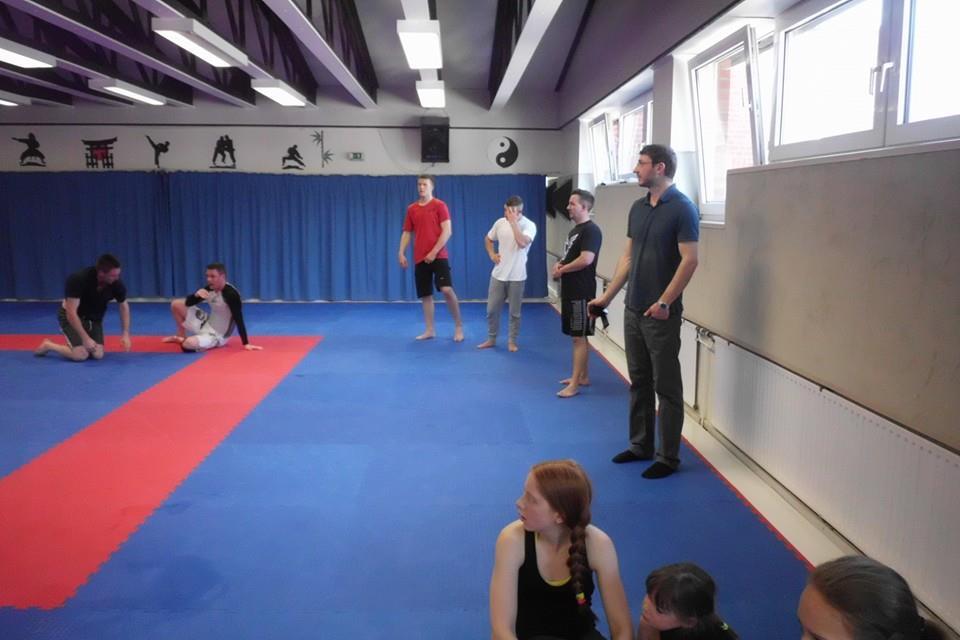 Gruppe von Menschen in einem Trainingsraum, einige sitzen auf blauen Matten, andere stehen um einen roten Bereich.