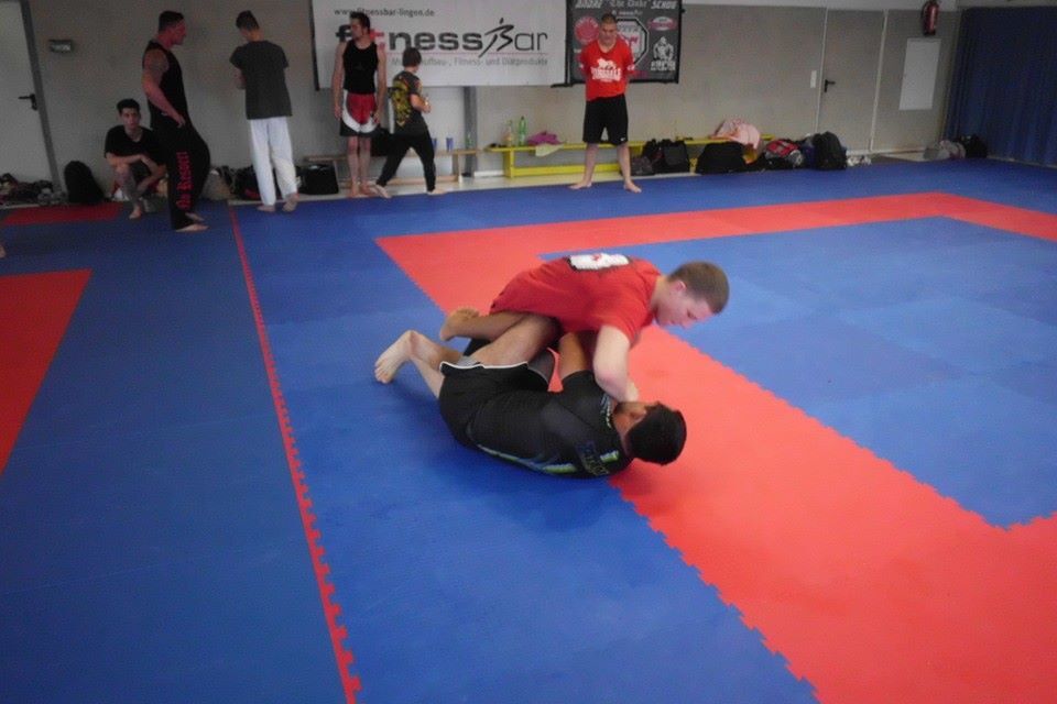 Zwei Kampfsportler &uuml;ben auf einer blauen Matte, einer sitzt auf dem anderen und kontrolliert ihn im Grappling.