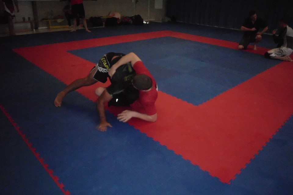 Zwei K&auml;mpfer in Sportkleidung k&auml;mpfen auf einem blauen und roten Kampfsportboden. Hintergrund mit weiteren Personen.