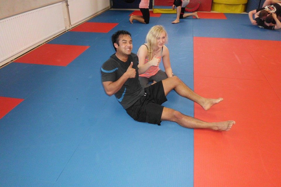 Zwei Personen sitzen auf mattenbedecktem Boden und zeigen Daumen nach oben, mit aktivem Training im Hintergrund.