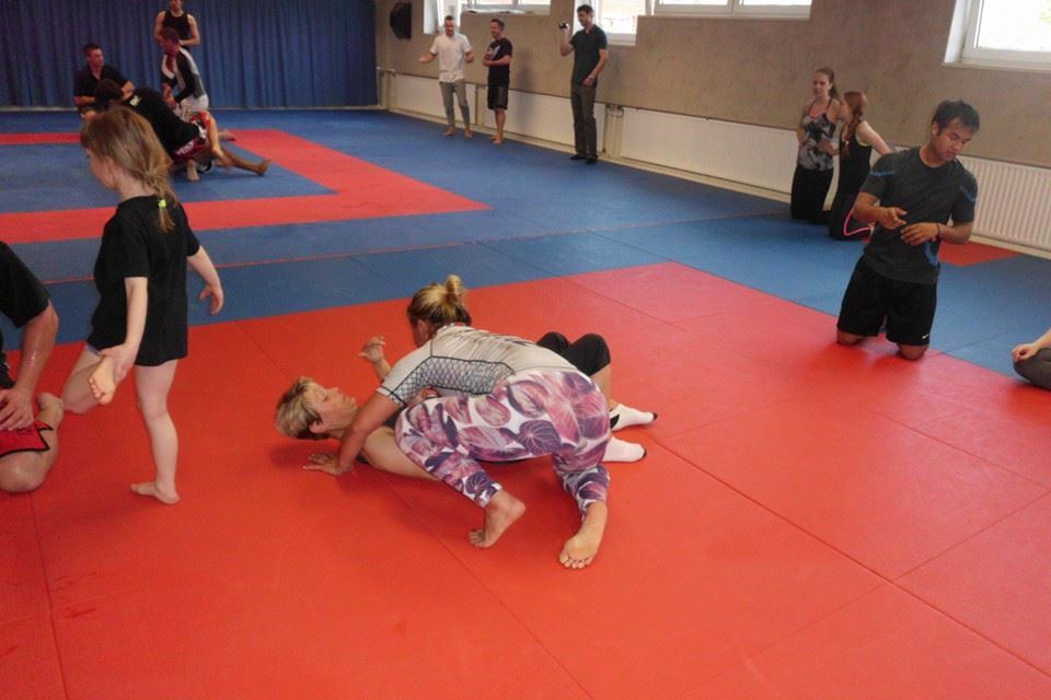 Zwei Personen trainieren Bodenkampf auf einer roten Mattenfl&auml;che, w&auml;hrend mehrere Zuschauer im Hintergrund stehen.