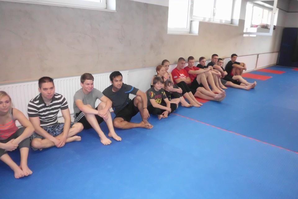 Gruppe junger Menschen sitzt auf einer blauen Matte in einem Trainingsraum, ans Wandsubstanz gelehnt.