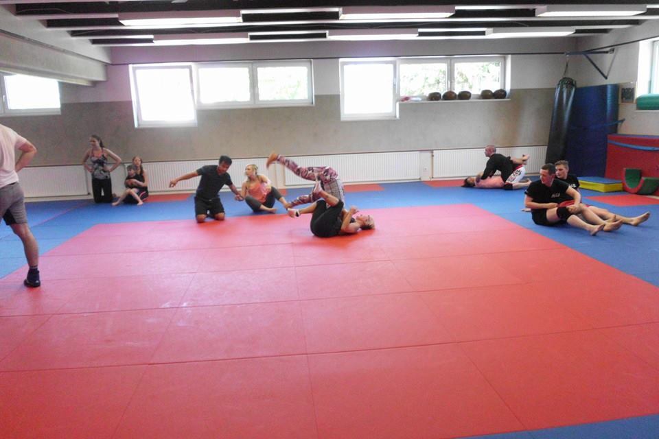 Gruppe von Menschen trainiert auf einer roten und blauen Matte in einem Kampfsportraum. Einige &uuml;ben Griffe, andere schauen zu.