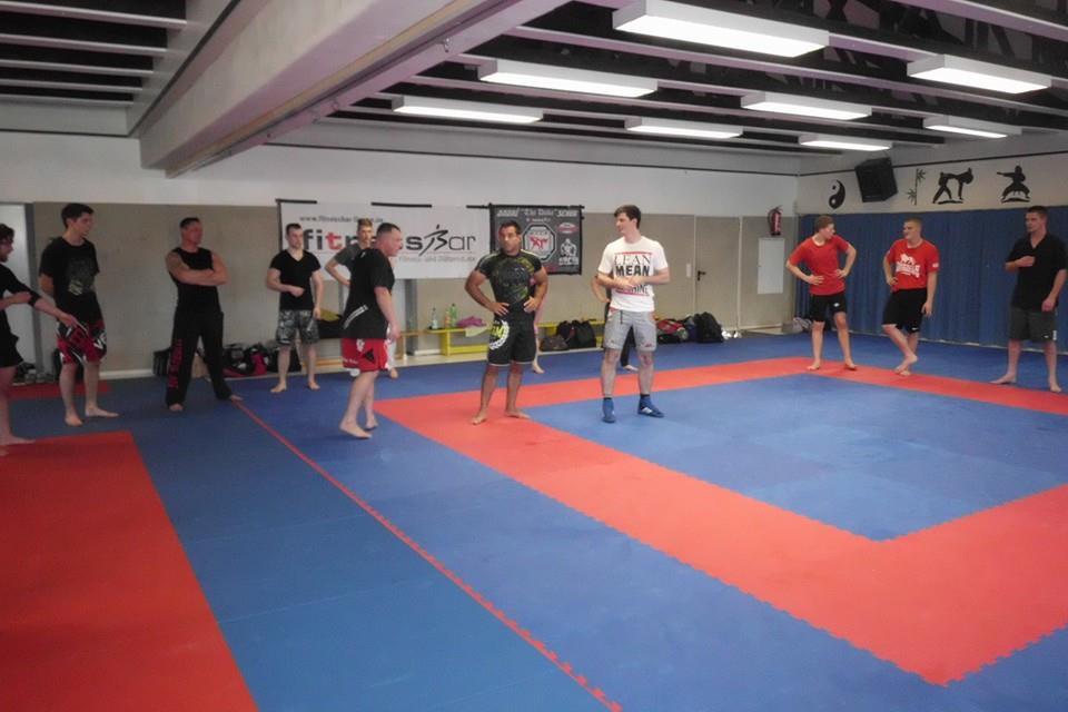 Gruppe von K&auml;mpfern in Trainingsanz&uuml;gen steht auf einer blauen Matte in einem Kampfsportstudio und bereitet sich auf &Uuml;bungen vor.