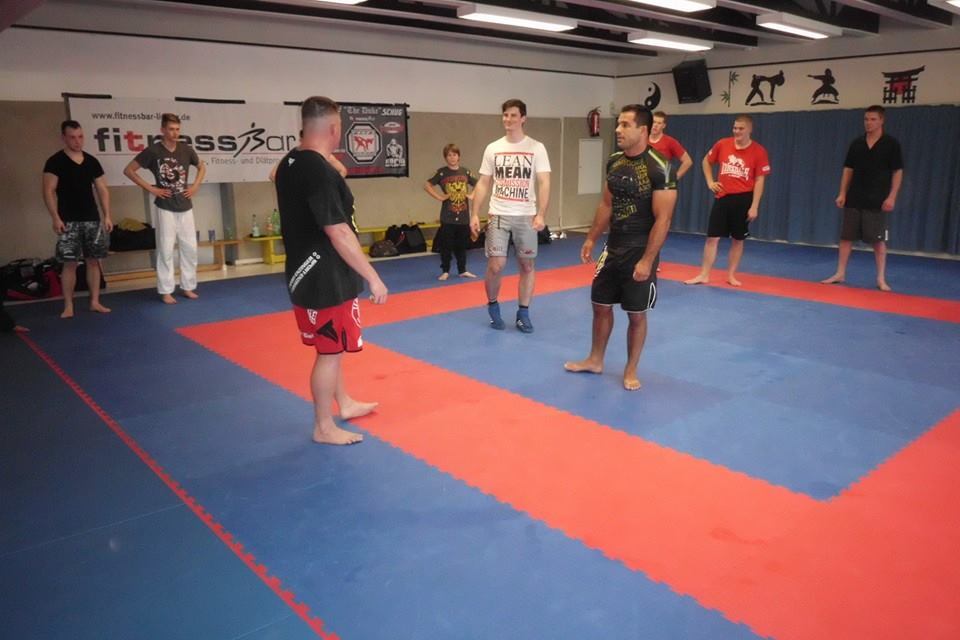 Sieben Personen stehen auf einer blauen und roten Trainingsmatte in einem Sportraum, bereit f&uuml;r eine Kampf&uuml;bung.