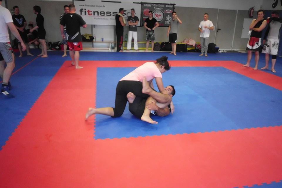 Zwei K&auml;mpfer trainieren auf einer Ringfl&auml;che, eine Person im rosa Shirt hat den Gegner in einer Griffposition.