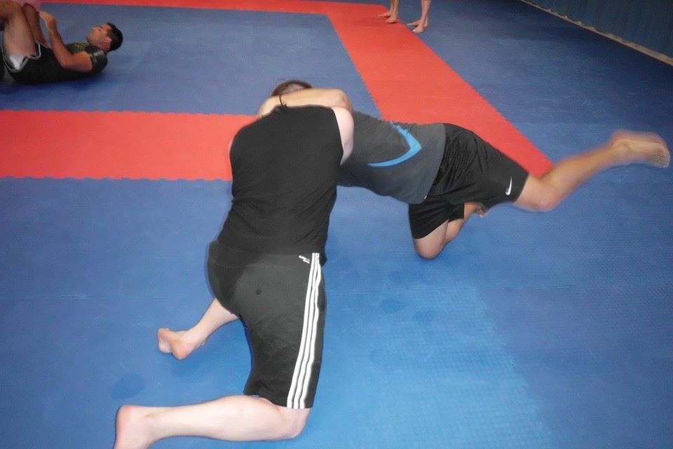 Zwei Sportler im Grappling halten sich auf einer blauen Matte, w&auml;hrend ein weiterer Sportler im Hintergrund &uuml;bt.