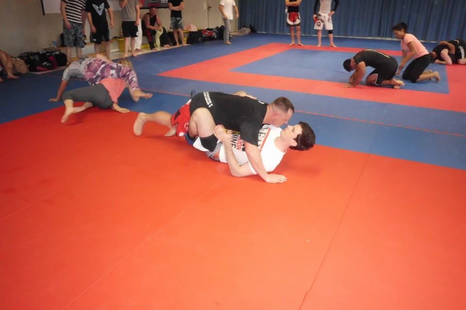 Zwei K&auml;mpfer &uuml;ben Grappling-Techniken auf einer roten und blauen Trainingsmatte, w&auml;hrend im Hintergrund weitere Teilnehmer trainieren.