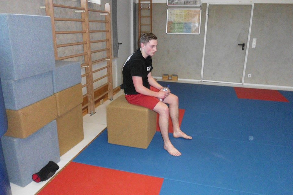 Junger Mann sitzt mit Wasserflasche auf einem quadratischen Block in einem Sportraum mit blauen und roten Matten.