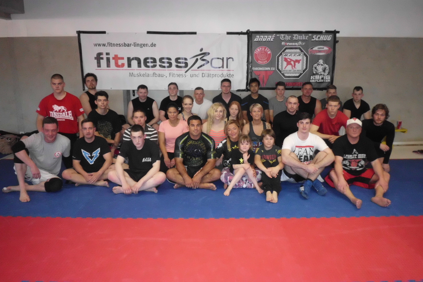 Gruppierung von 30 Personen in Sportkleidung, versammelt auf einer Matte mit Fitness-Werbung im Hintergrund.