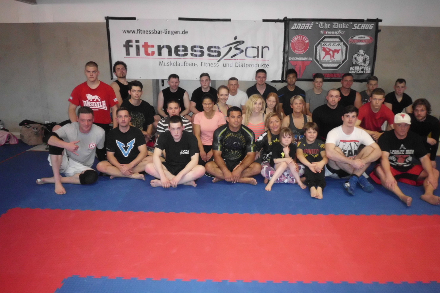 Gruppenaufnahme von Sportlern und Trainer auf einer bunten Matte in einem Fitnessstudio, mit Bannern im Hintergrund.