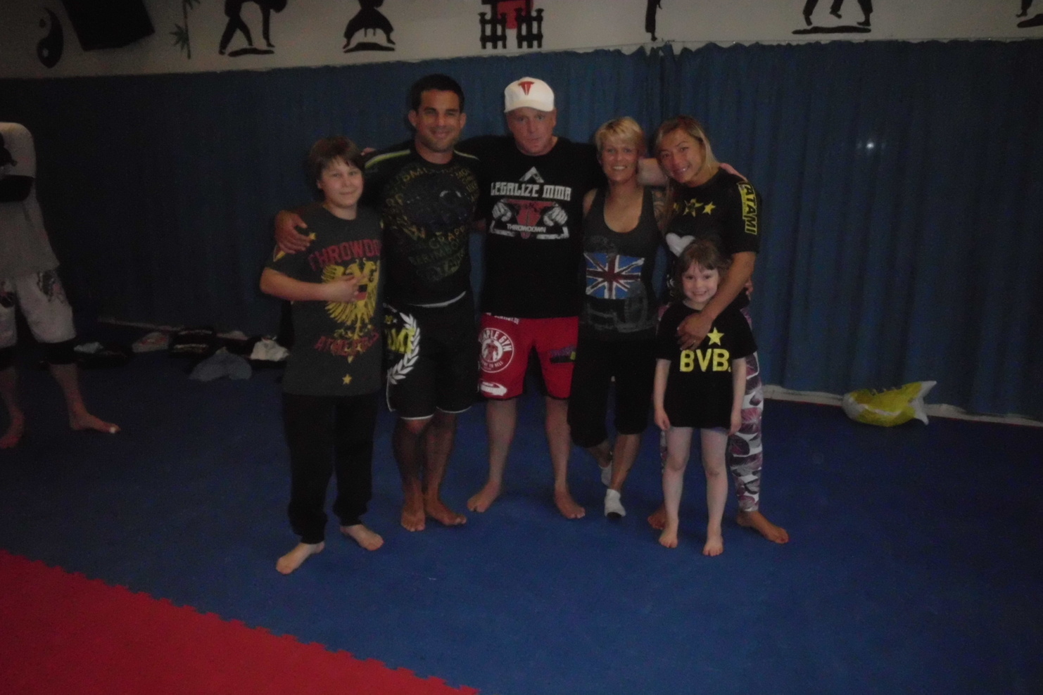 Familienfoto von f&uuml;nf Personen in Sportkleidung in einem Trainingsraum mit blauen Matten und einer blauen Wand.