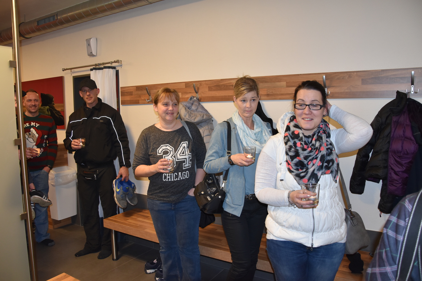 Gruppe von f&uuml;nf Personen steht in einem Raum, einige halten Gl&auml;ser, ein Kleiderst&auml;nder ist im Hintergrund sichtbar.