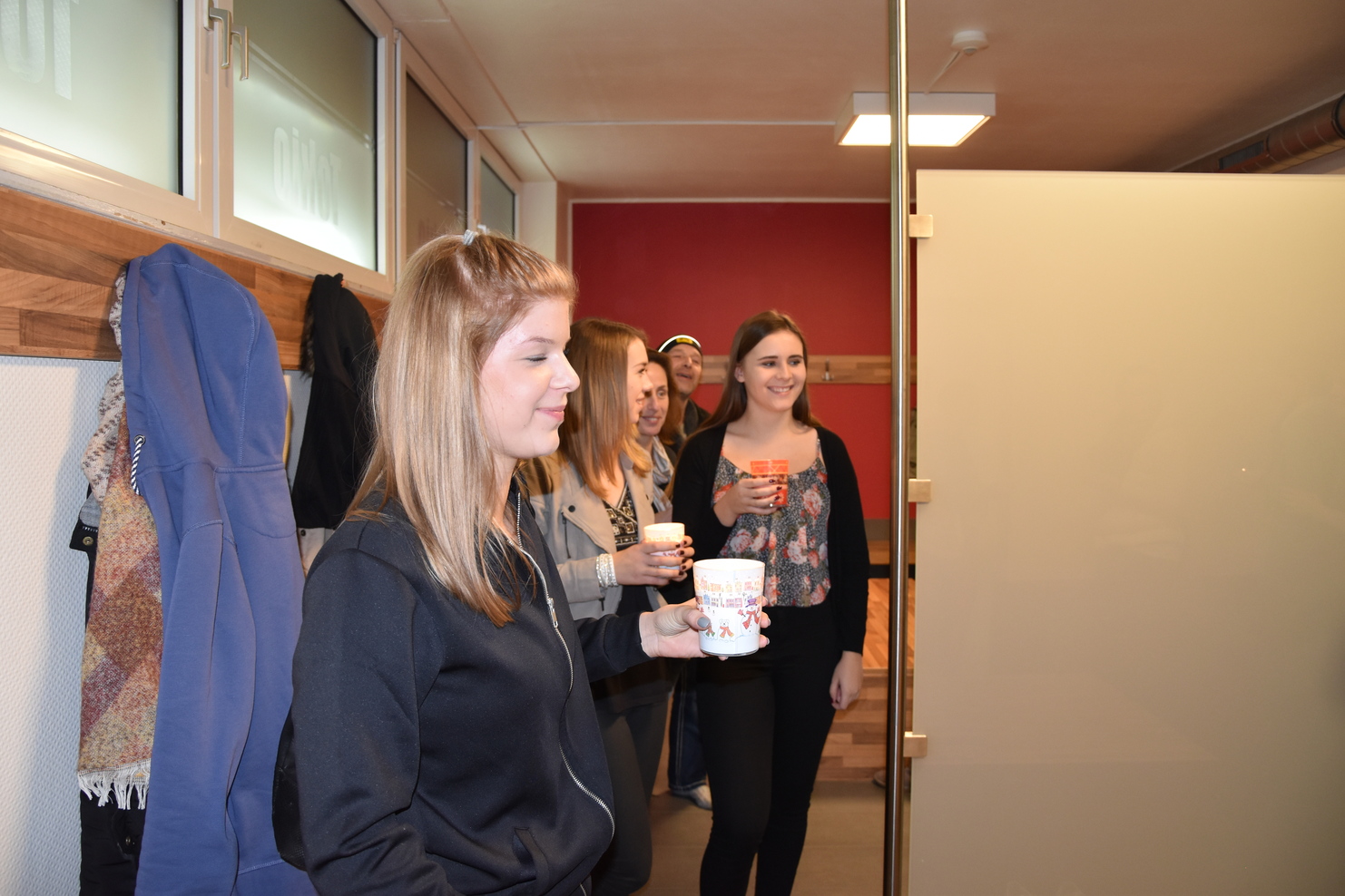 Vier Personen stehen in einem Raum, jede mit einem Becher in der Hand, vor einer transparenten Trennwand.