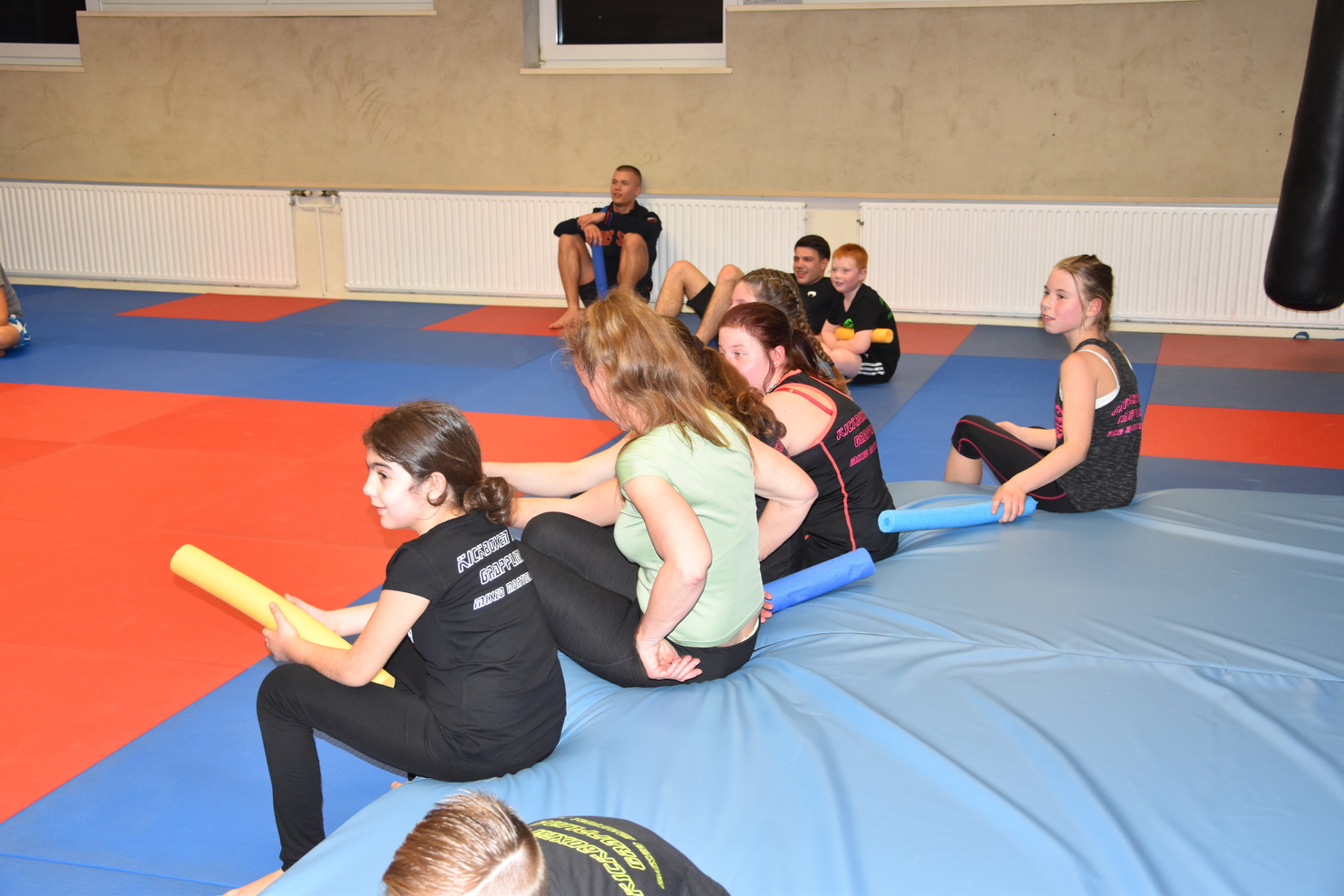 Kinder und Jugendliche sitzen auf einer blauen Matte im Trainingsraum, einige halten schaumstoffne Rohre in der Hand.