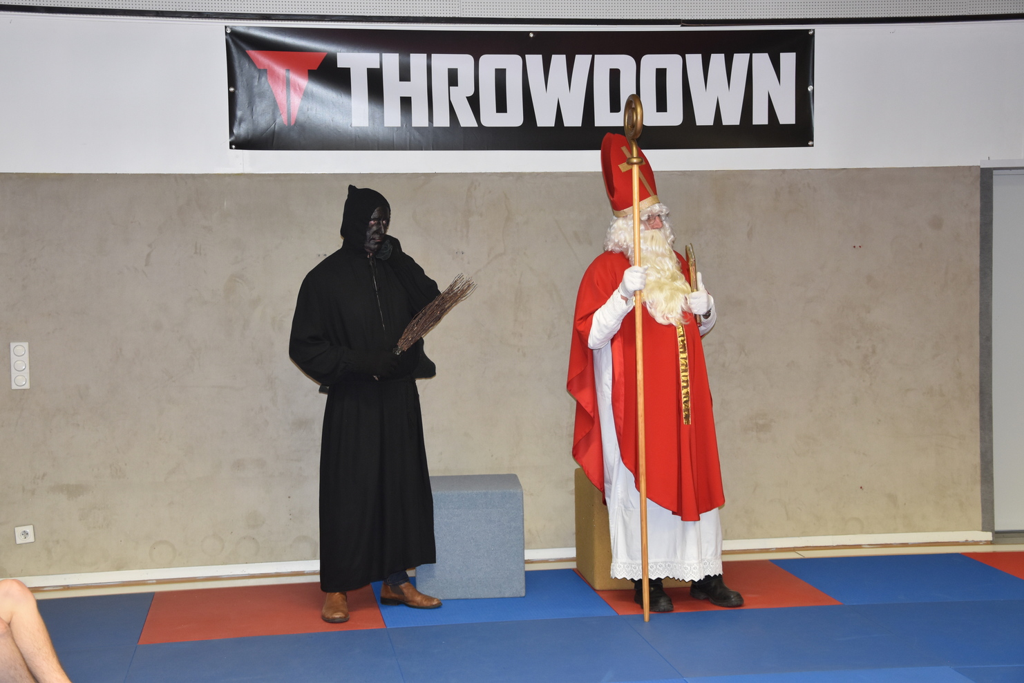 Zwei verkleidete Personen stehen in einer Sporthalle: ein roter Mantel tragender Nikolaus und eine schwarz gekleidete Gestalt.