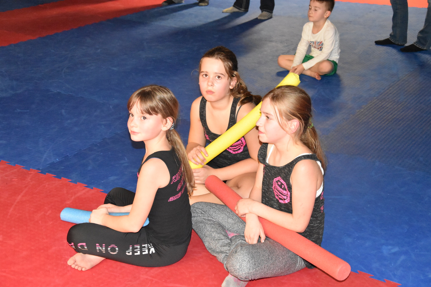 Drei M&auml;dchen in Sportkleidung sitzen auf einer Turnmatte und halten bunte Schaumstoffst&auml;be, w&auml;hrend ein Junge im Hintergrund spielt.