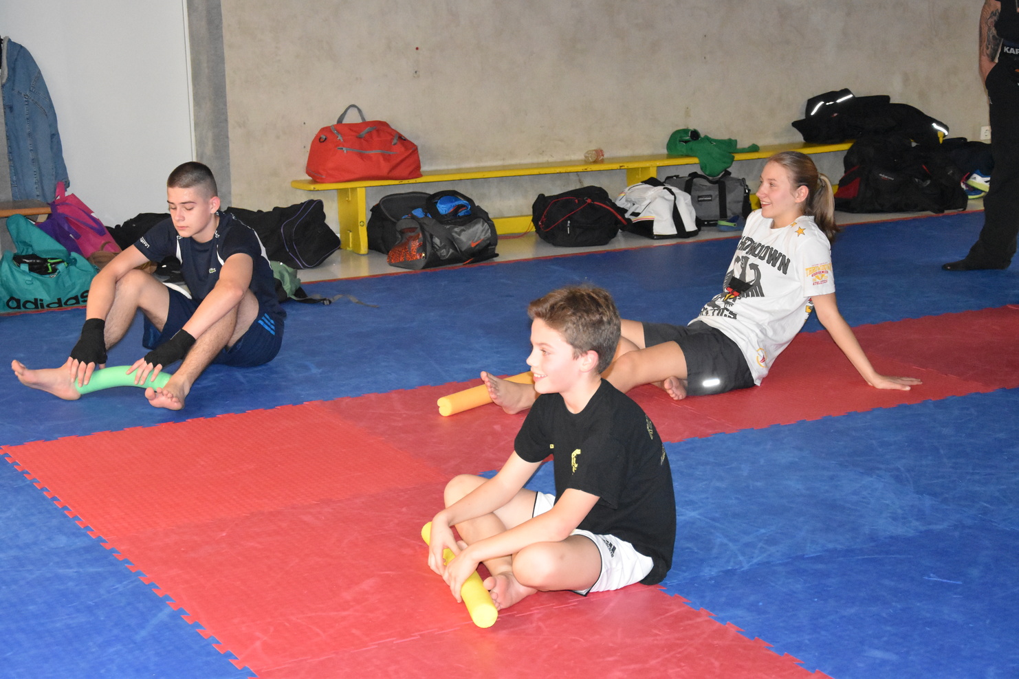 Drei Kinder sitzen auf Matten in einem Trainingsraum und dehnen mit gelben Schaumstoffrollen ihre Beine.