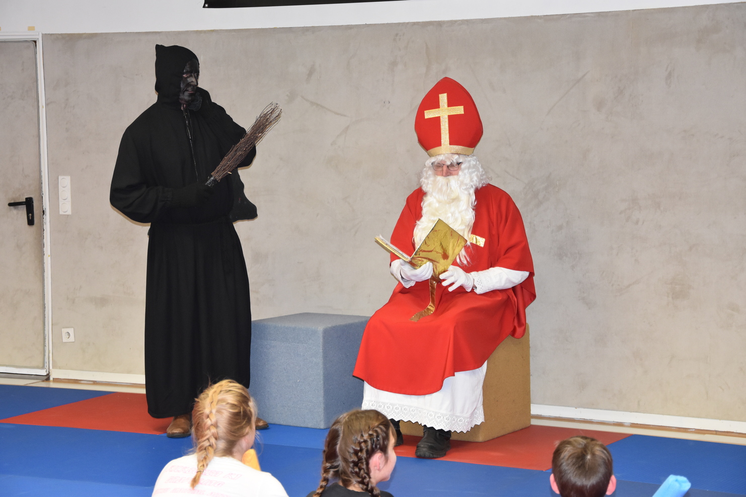 Nikolaus in rotem Gewand mit Buch sitzt auf einer Bank, w&auml;hrend eine dunkle Figur mit Besen daneben steht.