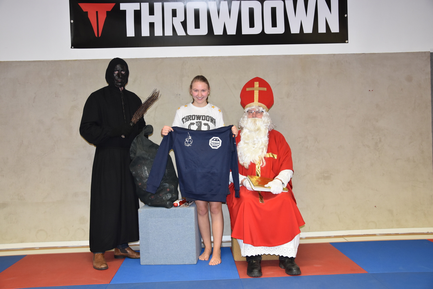 Ein M&auml;dchen in Sportkleidung h&auml;lt ein T-Shirt, umgeben von Santa und einer dunklen, mysteri&ouml;sen Figur.