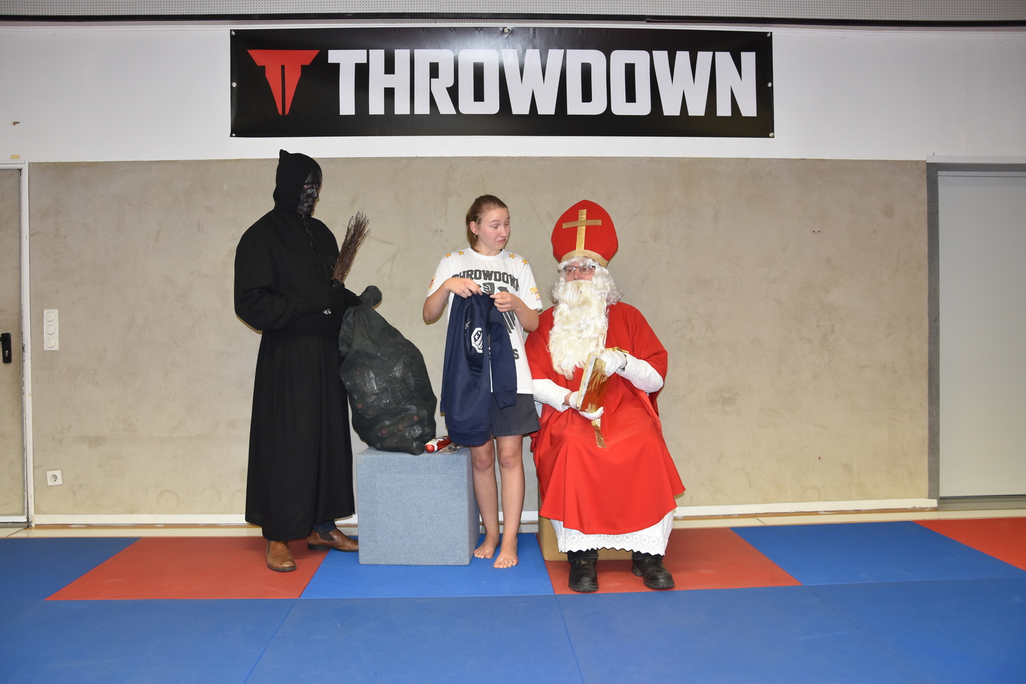 Kind mit Geschenkt&uuml;te steht neben einem Mann im schwarzen Kost&uuml;m und einem Weihnachtsmann in rotem Gewand auf einem blauen Boden.