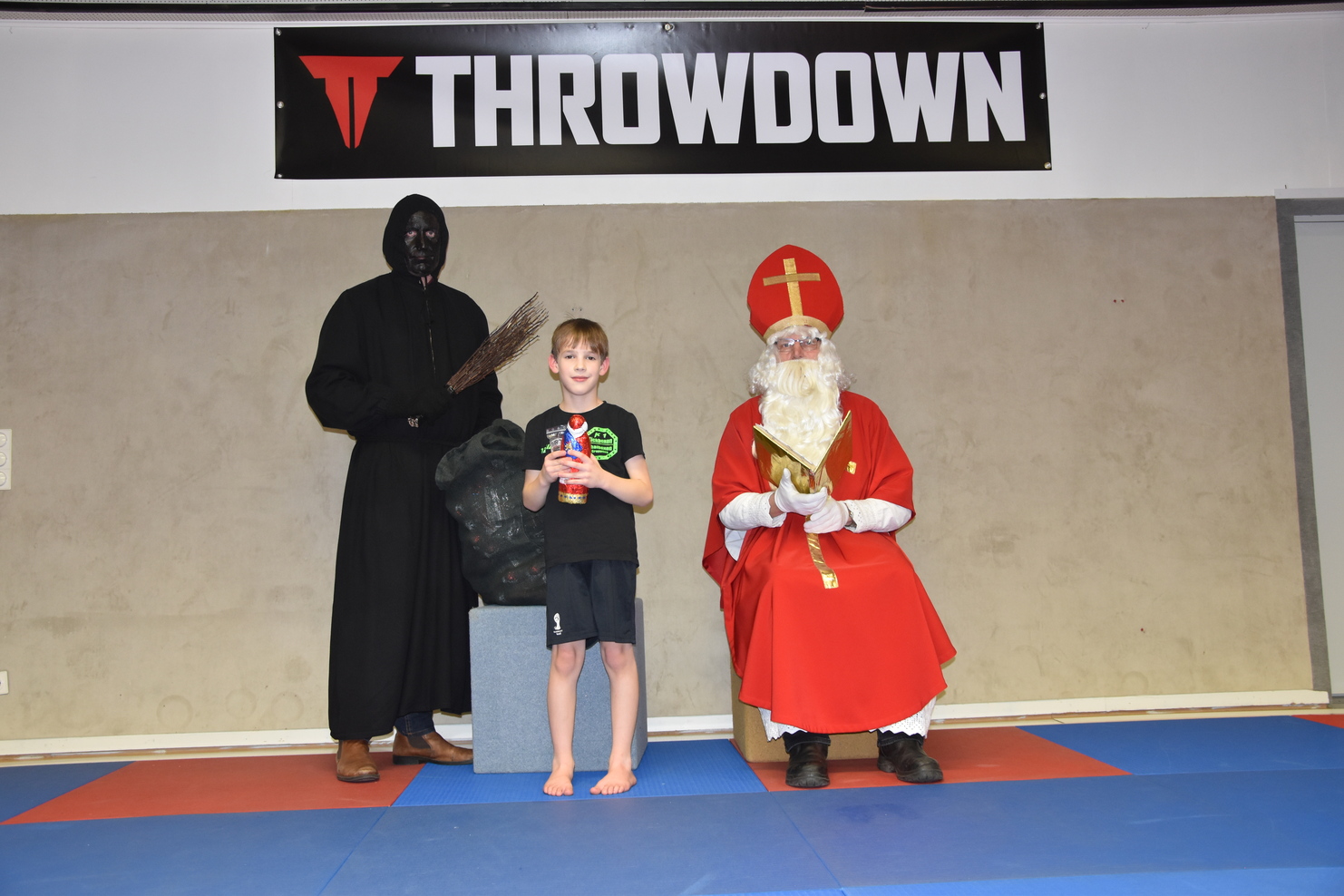 Junge mit Spielzeug steht zwischen einem finsteren персонажем und einem Weihnachtsmann in einer Sporthalle.