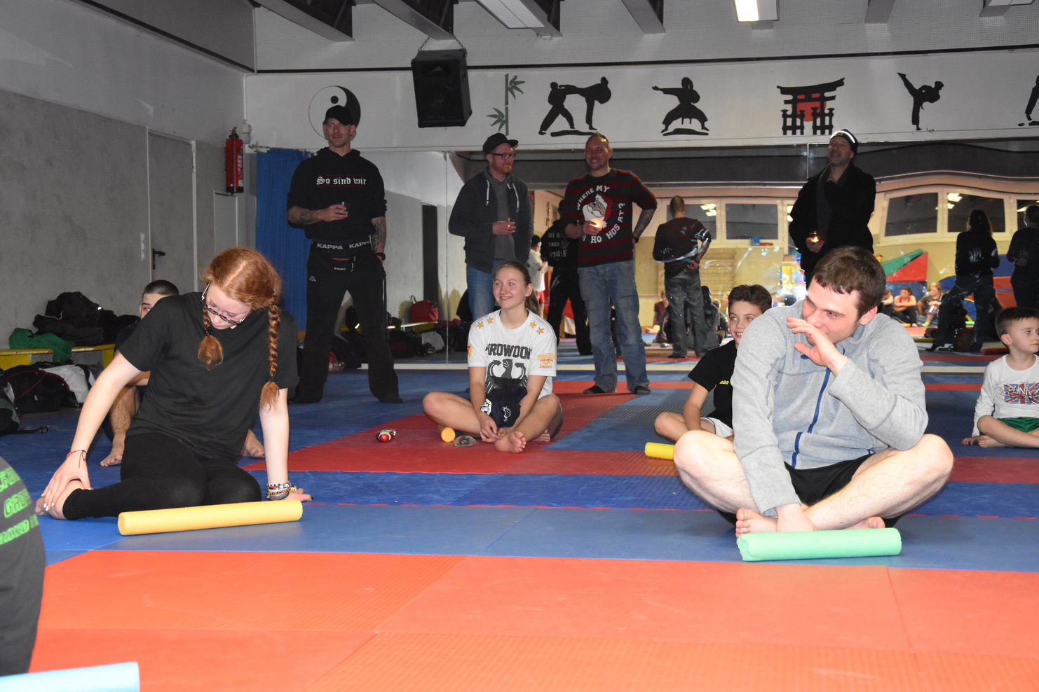Gruppenszene in einer Sporthalle: Teilnehmer verwenden Schaumstoffrollen zur Muskelentspannung auf einer bunten Matte.