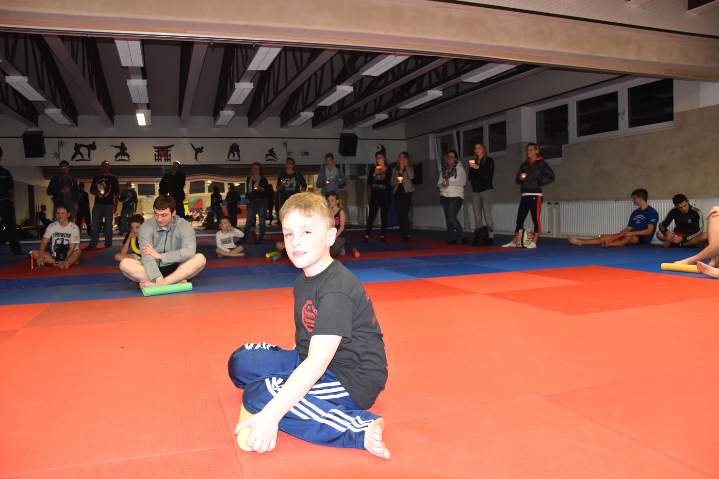 Junge sitzt auf der Matte in einem Sportraum, umgeben von Zuschauern, die ihm beim Training zuschauen.
