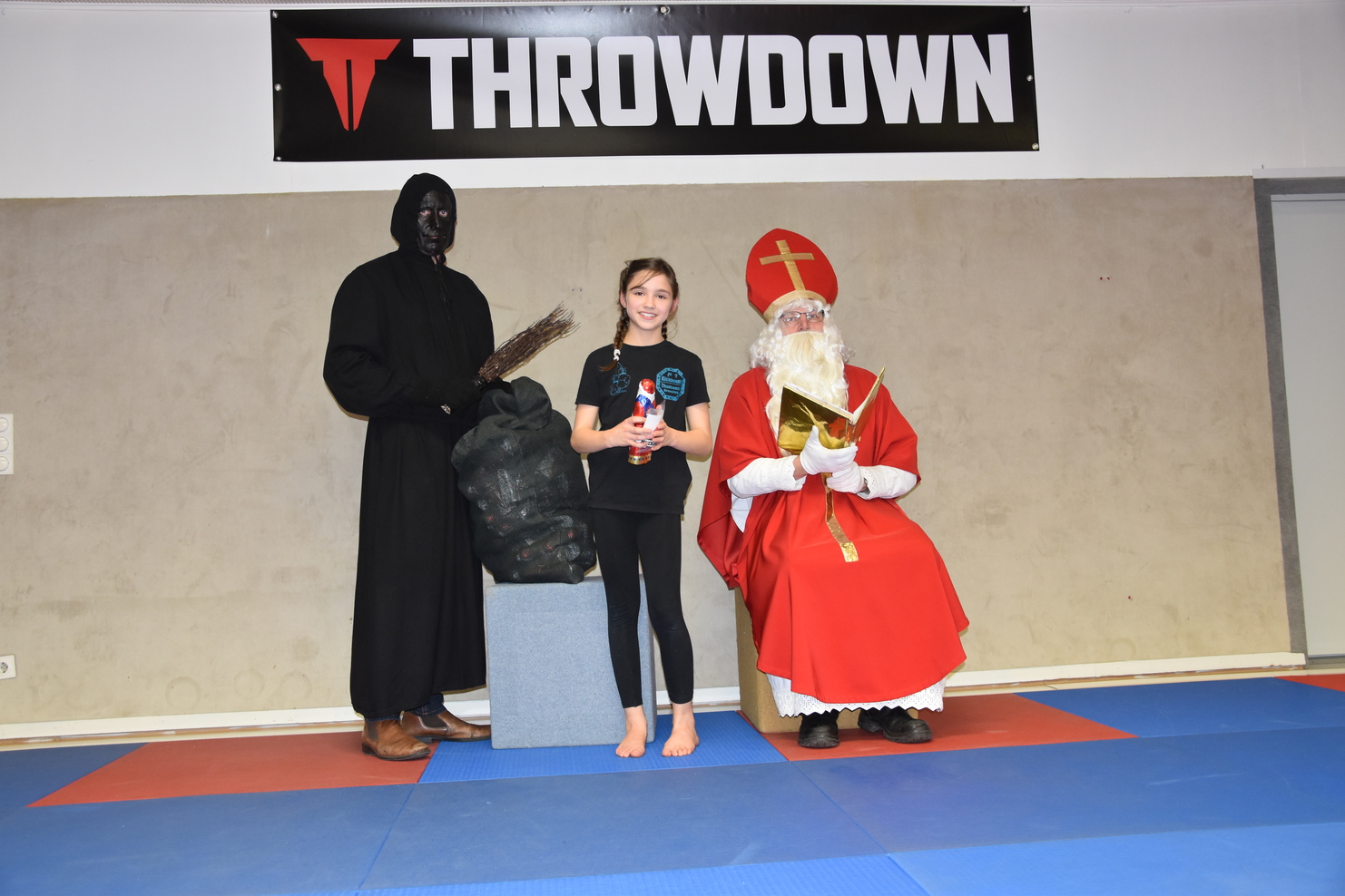 Ein Kind steht zwischen einem Mann im schwarzen Gewand und einem Weihnachtsmann auf einer Trainingsmatte.