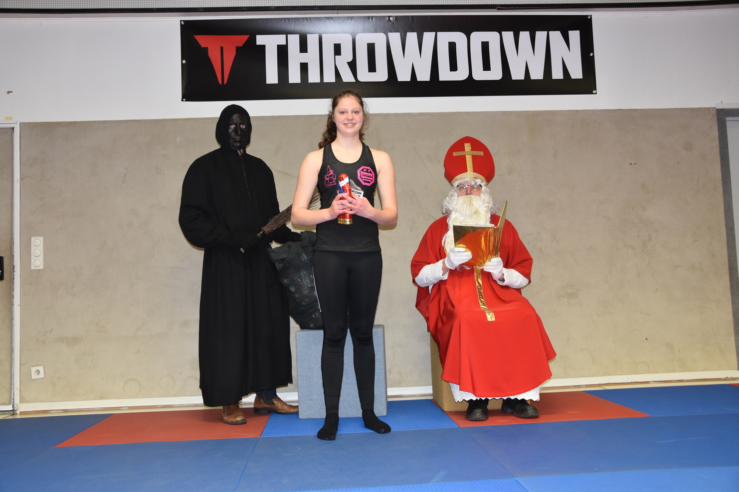 Eine Frau in Sportkleidung h&auml;lt eine Auszeichnung, umgeben von einem verkleideten schwarzen Charakter und einer Person in Rot.