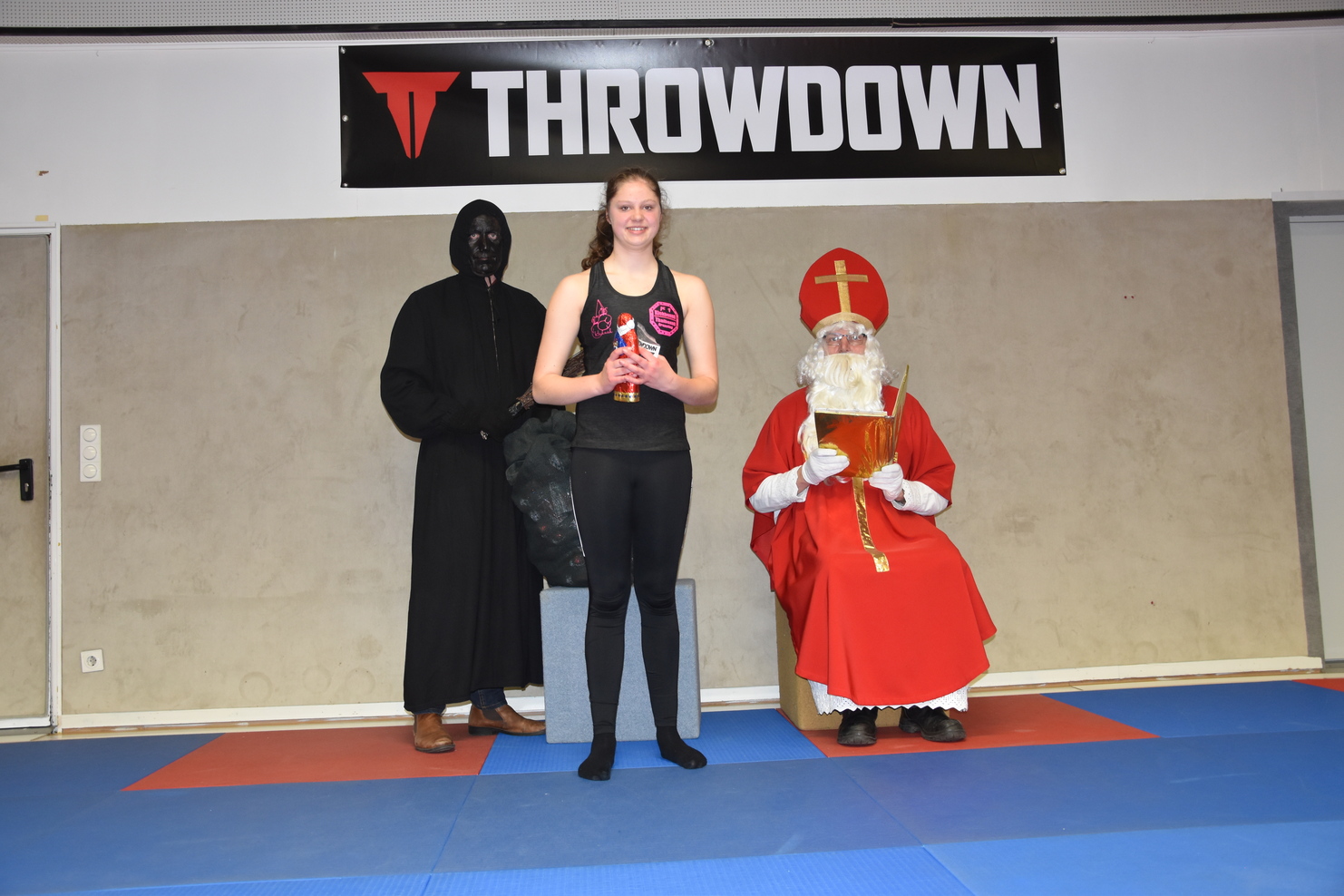 M&auml;dchen mit Pokal steht zwischen einem verkleideten Schatten und einem Weihnachtsmann in einer Sporthalle.