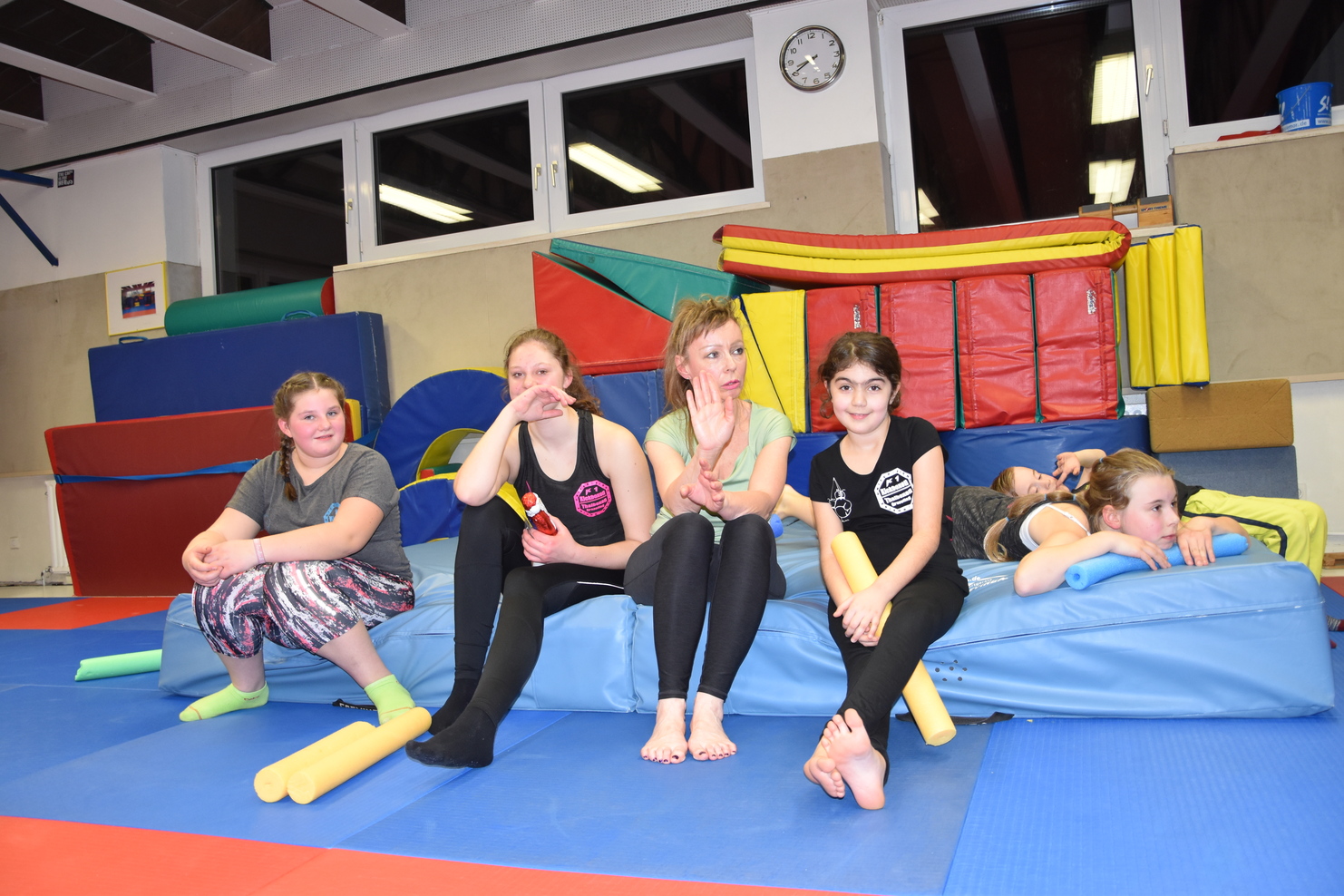 Sechs Jugendliche sitzen auf einer Gymnastikmatte in einem Sportraum, umgeben von Turnger&auml;ten und Schaumstoffbl&ouml;cken.