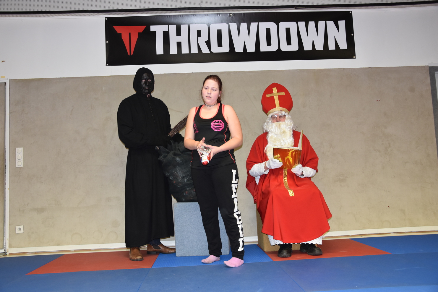 Drei Personen in ungew&ouml;hnlichen Kost&uuml;men stehen auf einer Trainingsmatte: ein schwarzer Umhang, eine Sportlerin und ein nikolaus&auml;hnlicher Mann.