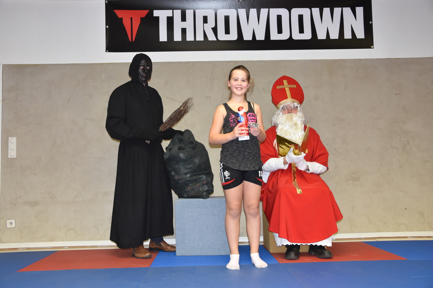 M&auml;dchen mit Getr&auml;nk steht zwischen einem schwarzen Kost&uuml;mtr&auml;ger und einem roten Weihnachtsmann in einem Sportraum.